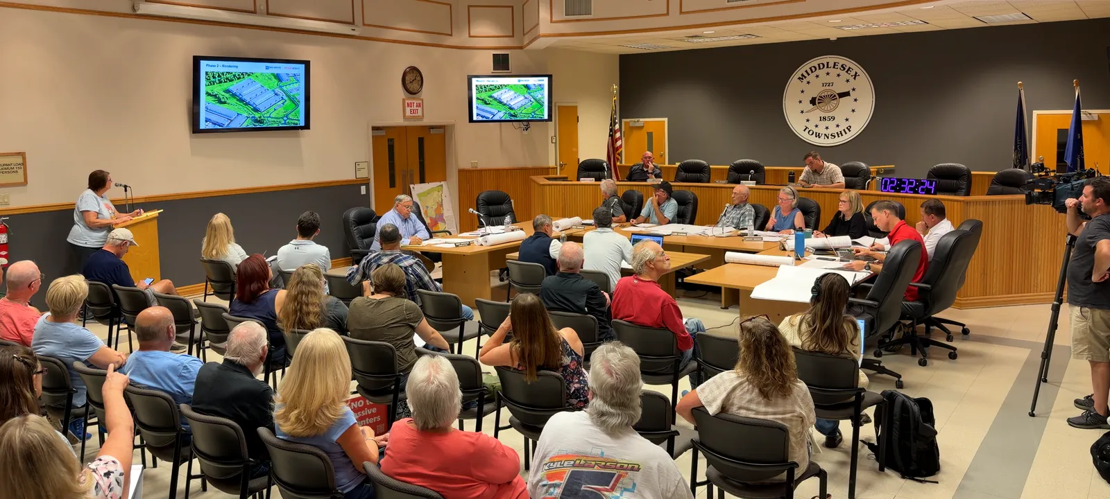 A full room of residents, seen from behind, at a Middlesex Township Planning Commission workshop. A resident speaks at the podium while the developer's Phase 2 rendering shows on the screens.