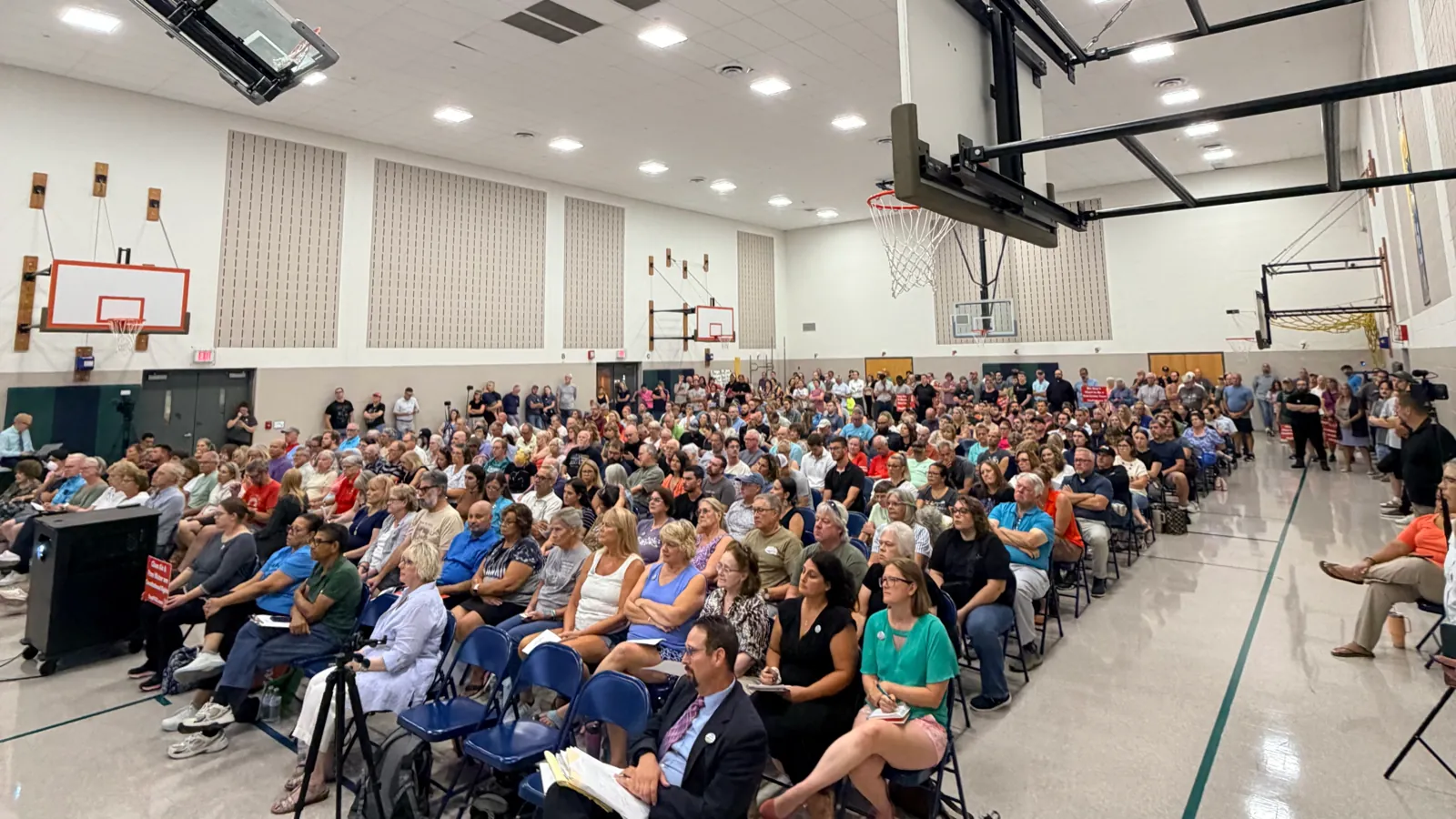A gym packed with residents at the Middlesex Township Board of Supervisors special meeting on August 19, 2026. Rows of neighbors fill the folding chairs, more stand along the back and side walls, and news cameras film from the side.
